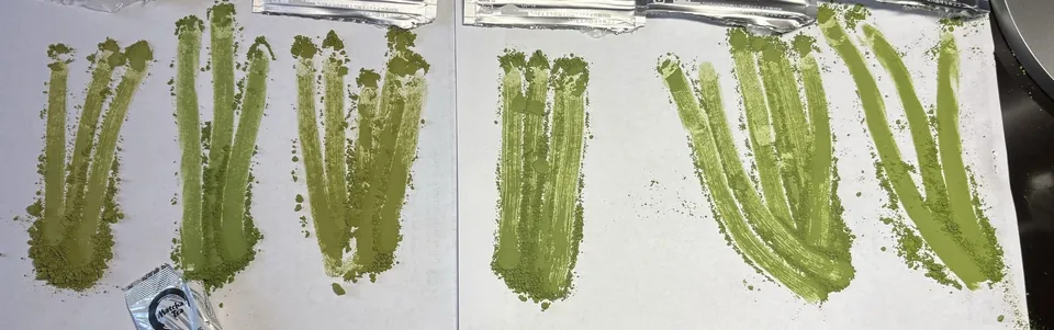 Šest vzorků matchy roztřených na bílém papíře, od olivově žluté po sytě zelenou